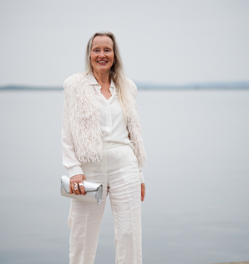 Sabine Weiskopf steht in ihrem weißen Hochzeitsanzug und Federjacke lächelnd am Ufer eines Sees